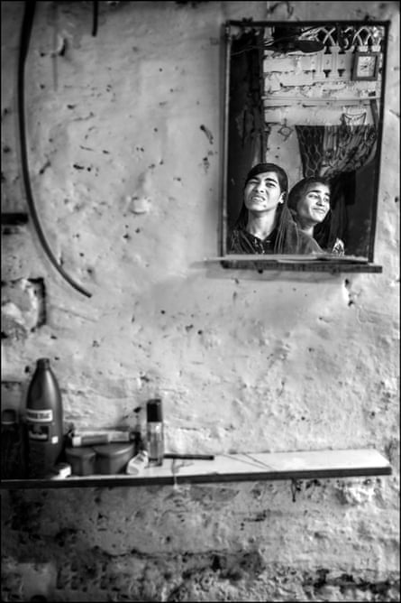 Two young women are seen reflected in a mirror on a crudely plastered wall. Both stare into space; one grimacing, the other smiling