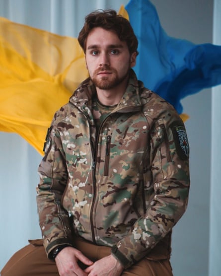 Young bearded man wearing army fatigues sits in front of a yellow and blue Ukrainian flag.