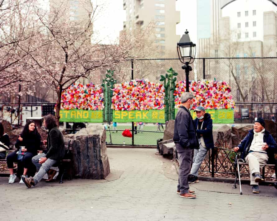 People sit on benches in a park, a colorful art installation on a fence in the background