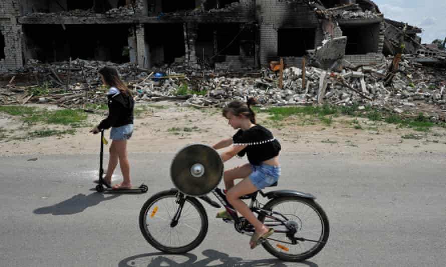 Children ride a bike and a scooter on a road in front of a destroyed building in the village of Novoselivka.