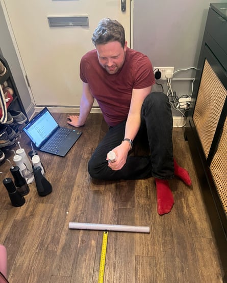 A man sits on a wooden floor holding a water flosser with a tape measure in front of him
