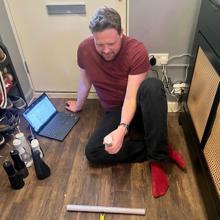 A man sits on a wooden floor holding a water flosser with a tape measure in front of him