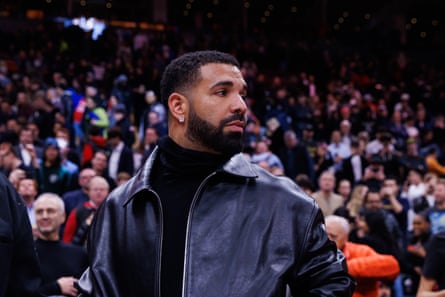 A bearded man with a leather jacket and black polo neck jumper standing in front of a crowd looking to his left