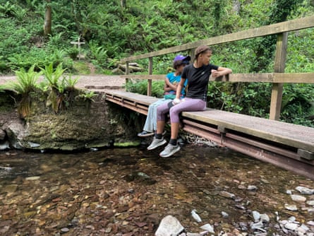 Two children sitting on a narrow wooden bridge over a stream.