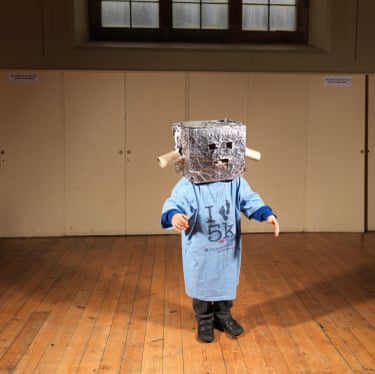 A child stands, arms stretched out in front, a silver-foil covered box on its head