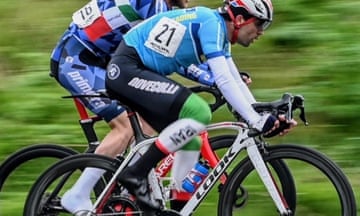 Mohammad Ganjkhanlou in a cycling race – he is slightly ahead of another rider whose head is not seen; he wears a light blue top with white long-sleeved underlayer and black shorts over green and white leggings; he rides a white bicycle