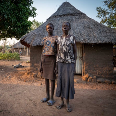 A tall, thin teenage African girl standing next to her mother outside a thatched hut 
