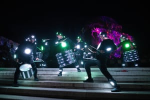 Drummers in black costumes that feature light displays; their drums also carry lights