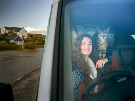 A woman sits at the wheel of a van with a tortoiseshell cat on her shoulder