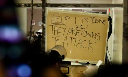Hands hold handmade sign that says “Help Us Please They Are Going to Attack” against a window in the dark.