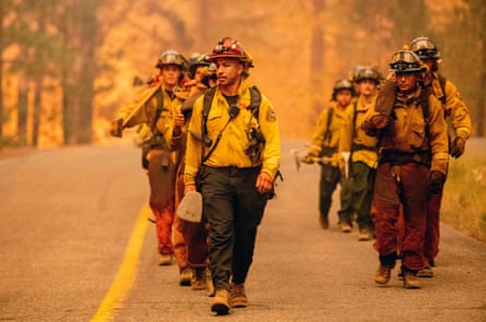 Firefighters in uniform walk down a road through smoky haze, with the sky between trees orange behind them.