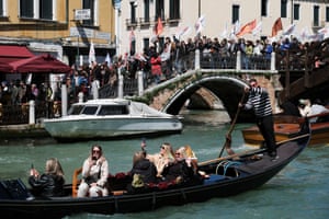 Tourists on a gondola taking photos as crowds of people stand on a bridge in the background waving flags