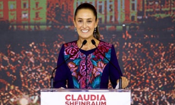A slim, middle-aged Latina woman with pulled-back brown hair and a color long-sleeved top in purple, blue, pink, and red, stands at a lectern that has her name on it, smiling.