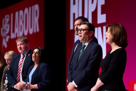 Andy Burnham, Lucy Powell, Shabana Mahmood and others in the background stand on stage singing.