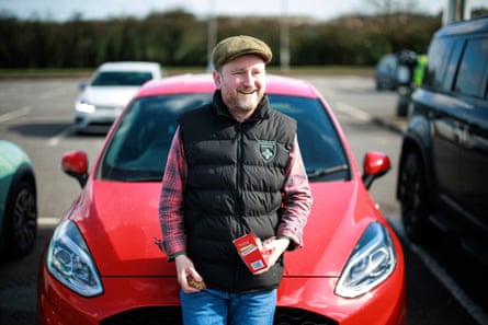 Roger Thorpe smiles and holds a boxed sanwich as he leans back on his car bonnet