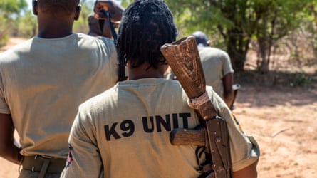 Rodah Chiawa trägt ein einfach aussehendes Gewehr in einem T-Shirt mit der Aufschrift „K9 Unit“ und zwei weiteren, alle von hinten gesehen.