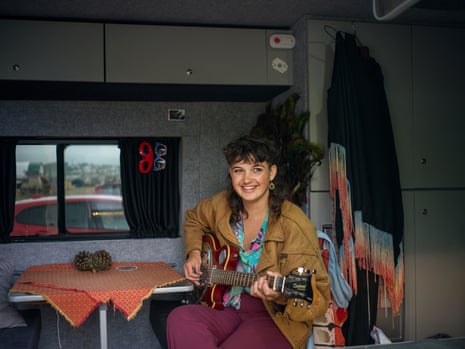 A young woman sits at a small table inside a van, holding a guitar.