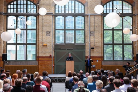 The view from the back of a seated audience watching Andy Burnham speak at a lectern in a high room with tall windows in a Victorian industrial building