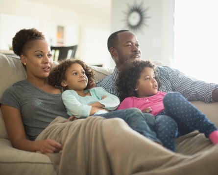 A family of two adults and two young children watch television while sitting close together on a sofa