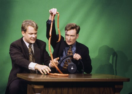 two men behind desk, with conan holding a string of sausages in one hand and a steering wheel in the other