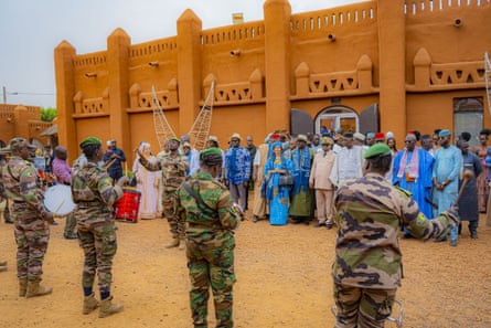 Musicians in camouflage fatigues play for a group of people in traditional west African robes outside a mudwalled-building
