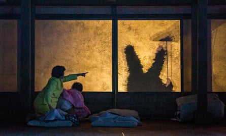 still image from stage performance of My Neighbour Totoro: two characters, one in yellow and the other in pink, are seen from behind looking through a window, which is lit in gold as the silhouette of a strange figure with long rabbit-like ears is seen through it, holding up a long, thin stick