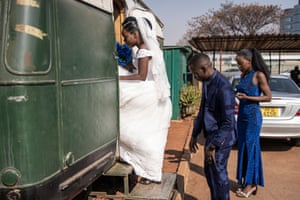 A woman wearing a white wedding dress steps into the caravan, holding a blue bouquet and followed by a man in a blue suit and a woman in a blue dress