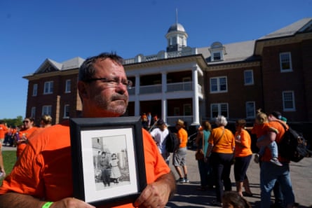 John Moses wearing an orange T-shirt stands outdoors holding a framed black-and-white photograph of his father