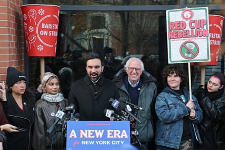 a man speaks into a microphone astatine a lectern arsenic group clasp signs adjacent him that publication ‘baristas connected strike’ and ‘red cup rebellion’