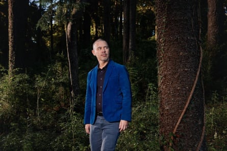 A man in a blue jacket, standing against a backdrop of trees.