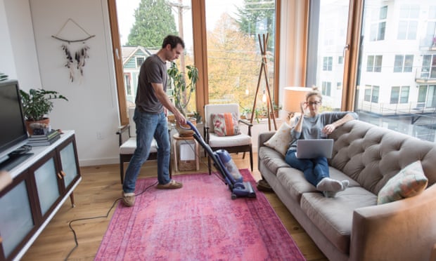 A man is vacuuming while a woman is reclining on the sofa with a laptop