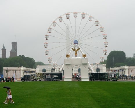 People under an umbrella cross a lawn in front of a Ferris wheel