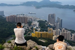 Two people sit on top of rocky ledge as they point cameras towards tall buildings next to the sea as a large vessel traverses the water