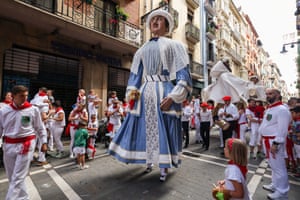 A giant effigy of a woman makes its way down a street followed by a marching band and surrounded by people in white costumes with red neckties and sashes