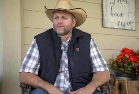 A man wearing a short-sleeved plaid shirt, black vest and tan cowboy hat sits outside while looking off to the side.