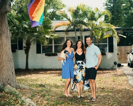 Kasey, Rachael and Aaron standing in their front garden. Kasey is holding a small dog. Part of an LGBTQ+ flag can be seen hanging from a tree