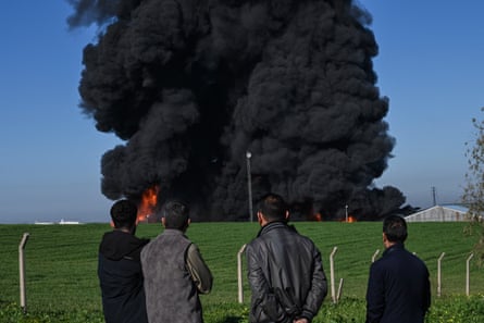 Four people watch from a safe distance as thick black smoke billows up from an oil warehouse