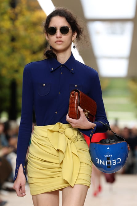 A model on the catwalk wearing a deep blue shirt with pale yellow short wrap skirt and carrying a blue cycle helmet with the logo Celine.