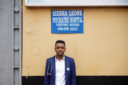 A young doctor with a stethoscope around his neck stands outside beside a painted wall with a hospital sign behind him.