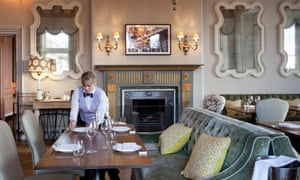 A waitress in a bow-tie setting a table with cushioned sofa seating and a gilded fireplace behind