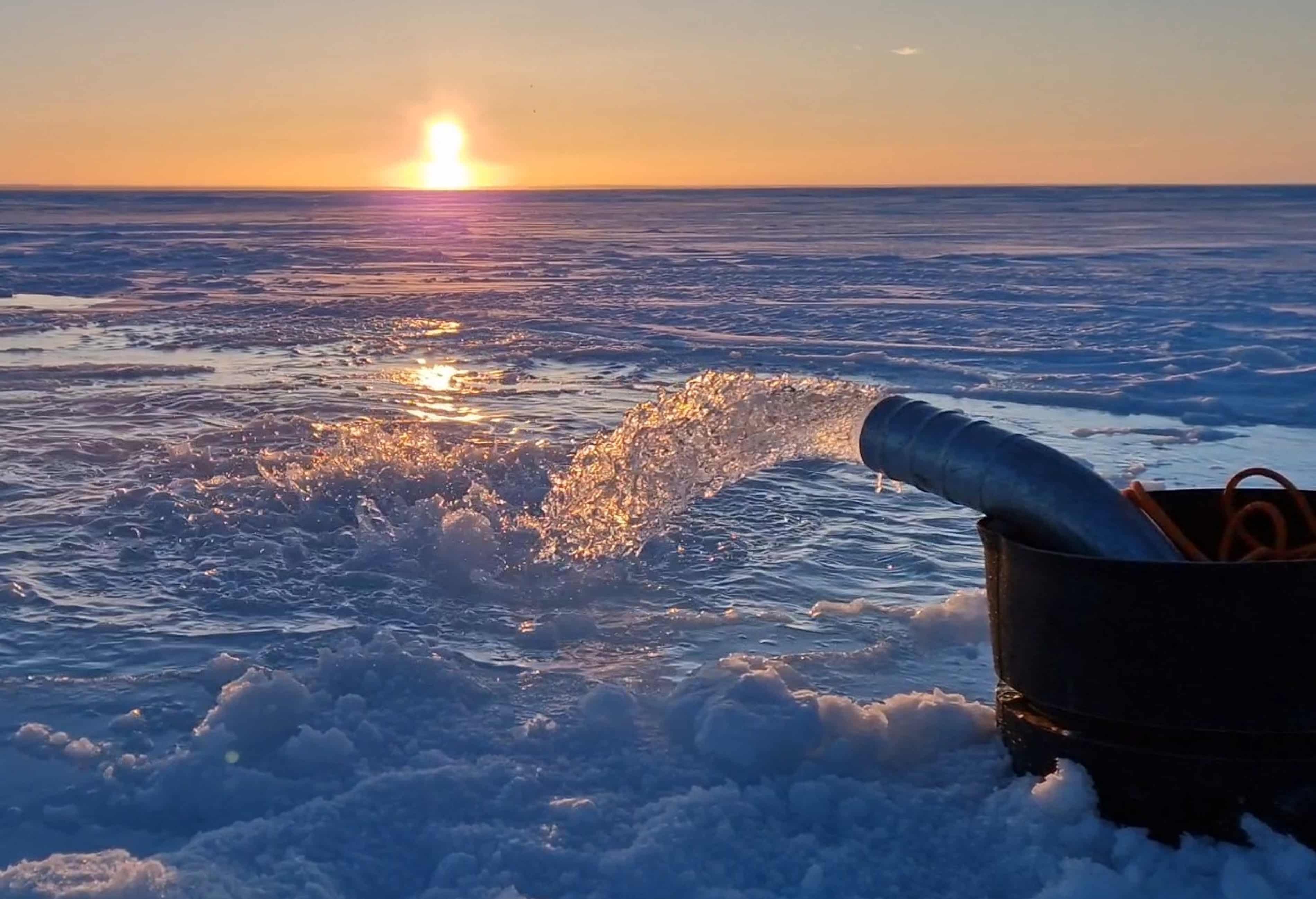 Tubo metálico expulsando agua sobre una superficie helada en plena puesta de sol, con reflejos dorados en el hielo, posiblemente en un experimento científico en el Ártico o la Antártida.
