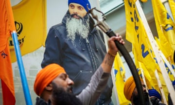 A turbaned man holds a sword in front of a poster featuring the photo of another turbaned man.