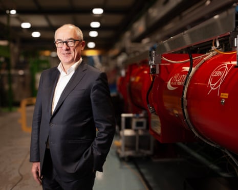 Mark Thomson smiling in suit next to red machine with Cern logo on it