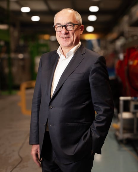 Mark Thomson smiling in suit next to red machine with Cern logo on it