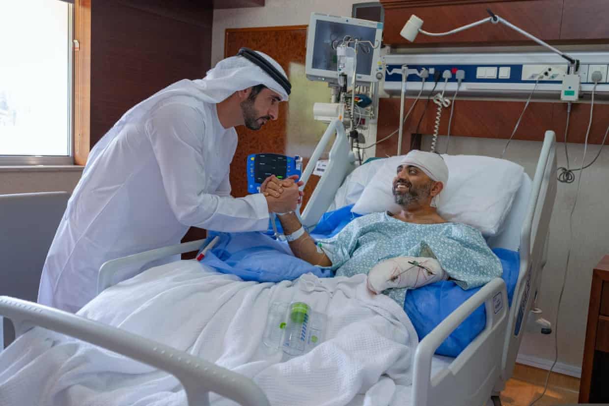 The crown prince, wearing a white robe and headdress, leans over the hospital bed to shake hands with the pilot, who wears a pale green hospital gown and has his head and one hand and forearm heavily bandaged. Both men are smiling.