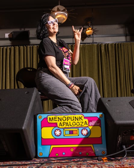 A woman smiles and throws up the sign of the horns while posing next to a sign that says ‘Menopunkapalooza’