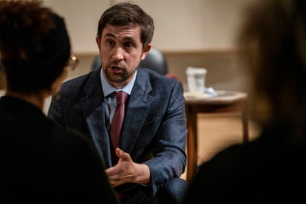 A young white man with brown hair and a trim beard wearing a suit leans in, speaking and gesturing, as he appears to talk to two women silhouetted in the foreground.