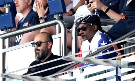 Snoop Dogg sat in the stands wearing a black cap and a stars and stripes jacket.