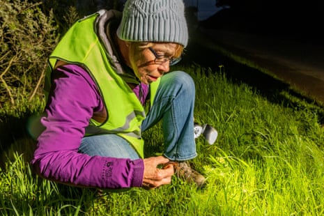 A woman wearing a purple jacket and green vest kneels and holds a newt