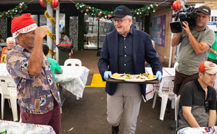 The prime minister holds a tray of food and is greeted by a guest wearing a Santa hat